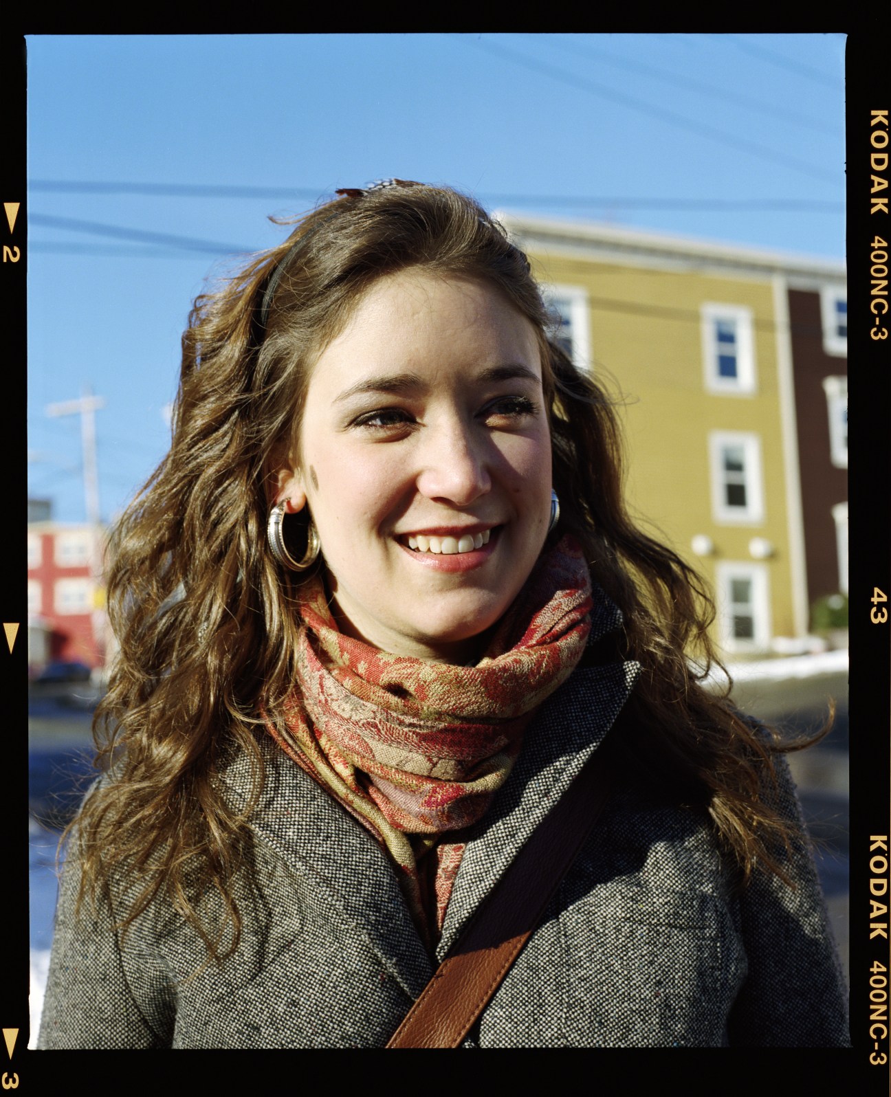 A smiling woman with wavy brown hair wearing a grey jacket and patterned scarf stands in front of colorful buildings in St John's.