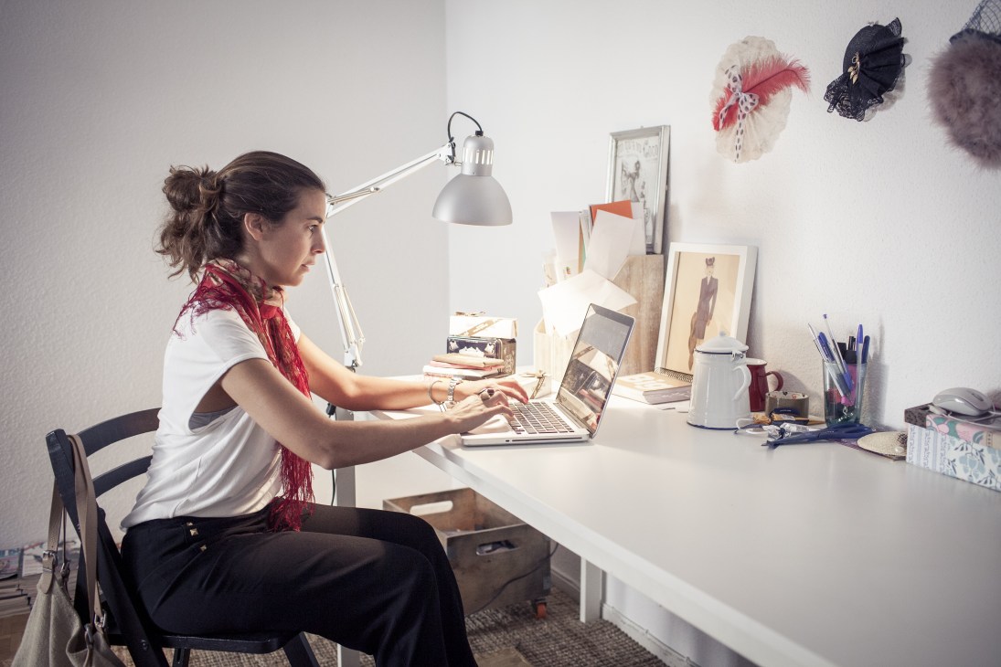 Rocio Muñoz of Real Fábrica Española works at her laptop in a bright workspace decorated with fashion sketches and accessories.