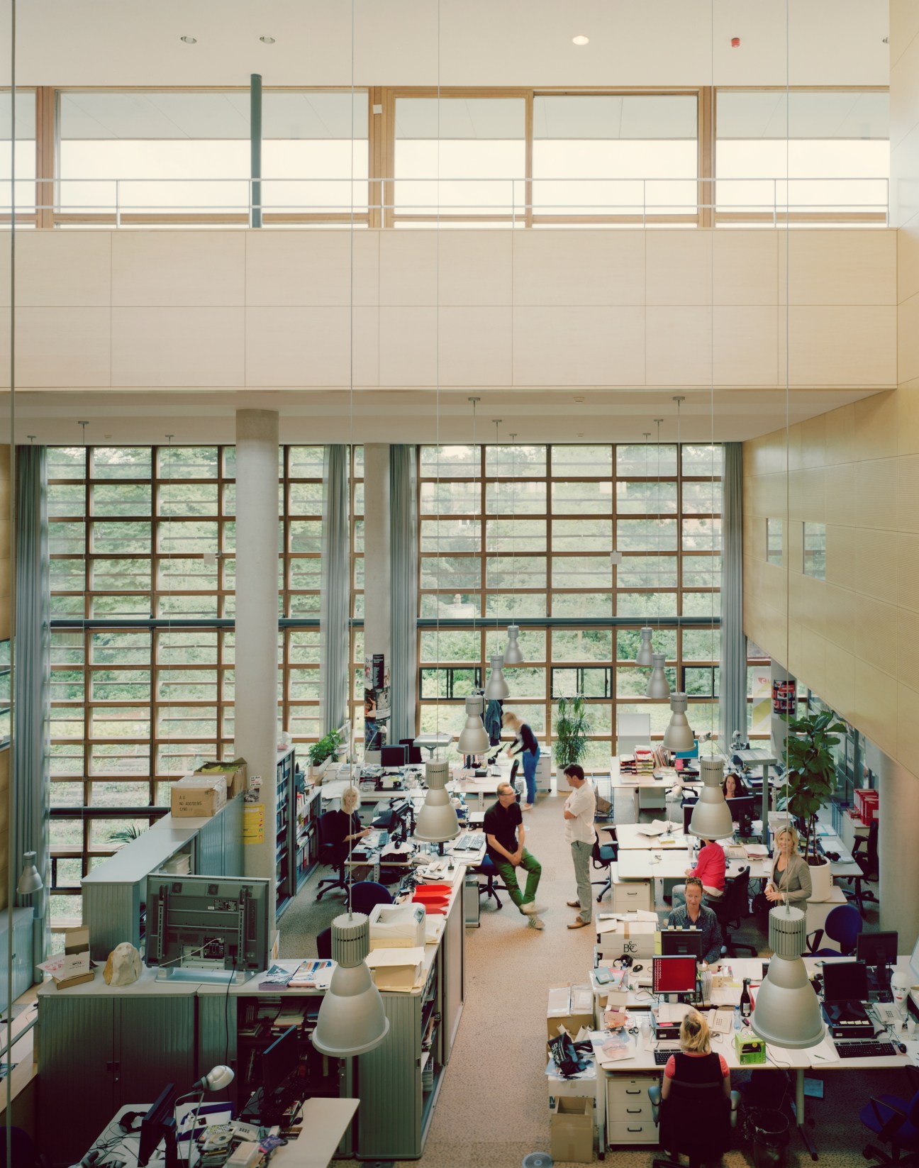 Modern open-plan office with high ceilings, large windows overlooking greenery, and staff working at desks throughout the space.