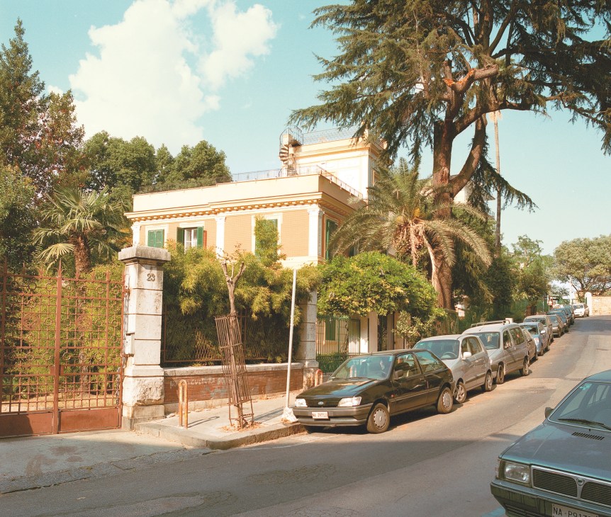 A tree-lined street in Naples with a peachy-orange neoclassical villa and parked cars along the road.