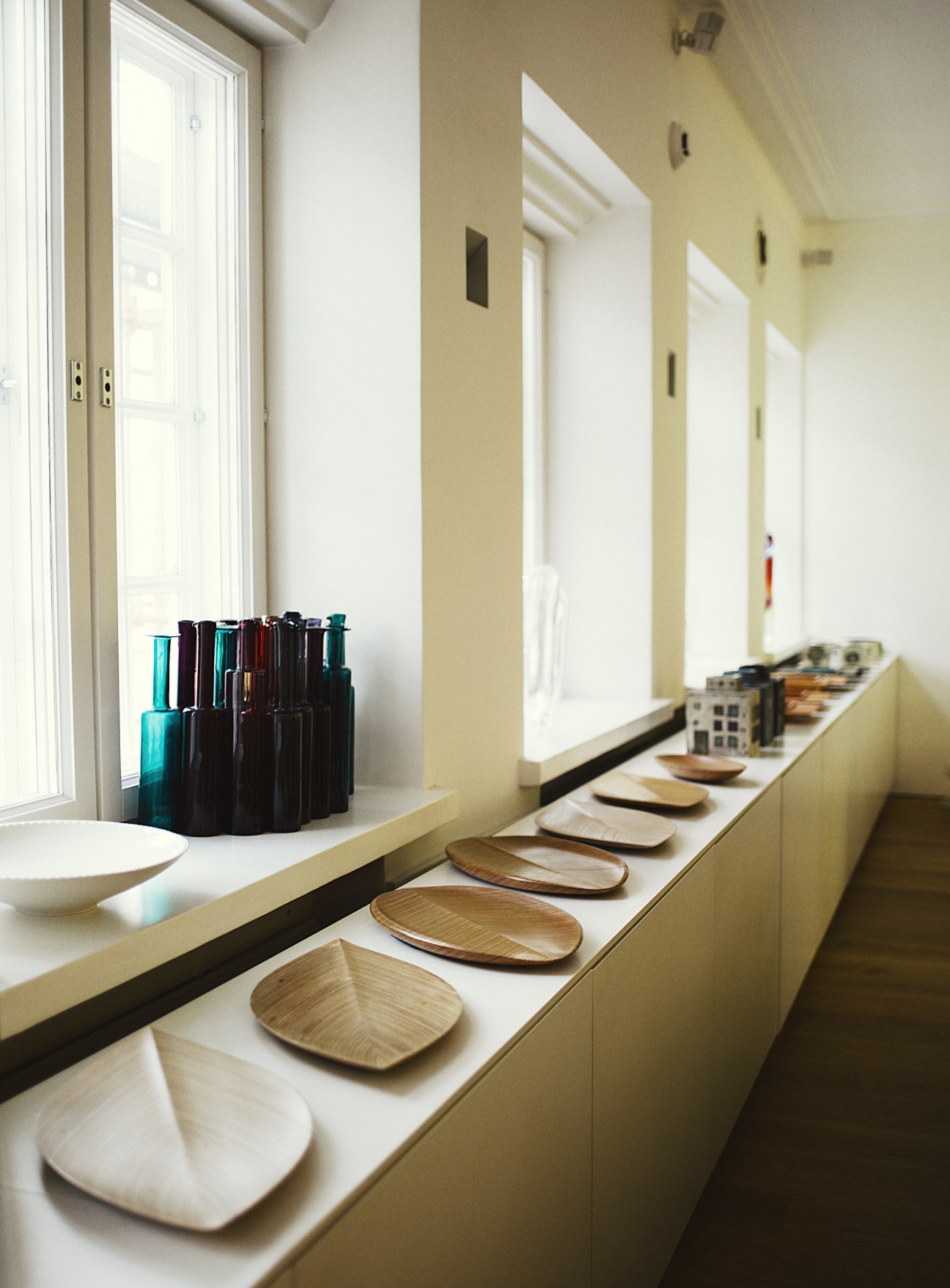 Kitchen counter displaying plywood plates by Tapio Wirkkala, glass bottles by Nanny Still, and ceramic pieces by Rut Bryk along sunlit windows.