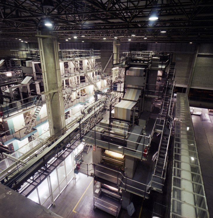 Interior of a large newspaper printing plant with industrial printing machinery, metal catwalks, and overhead steel framework.