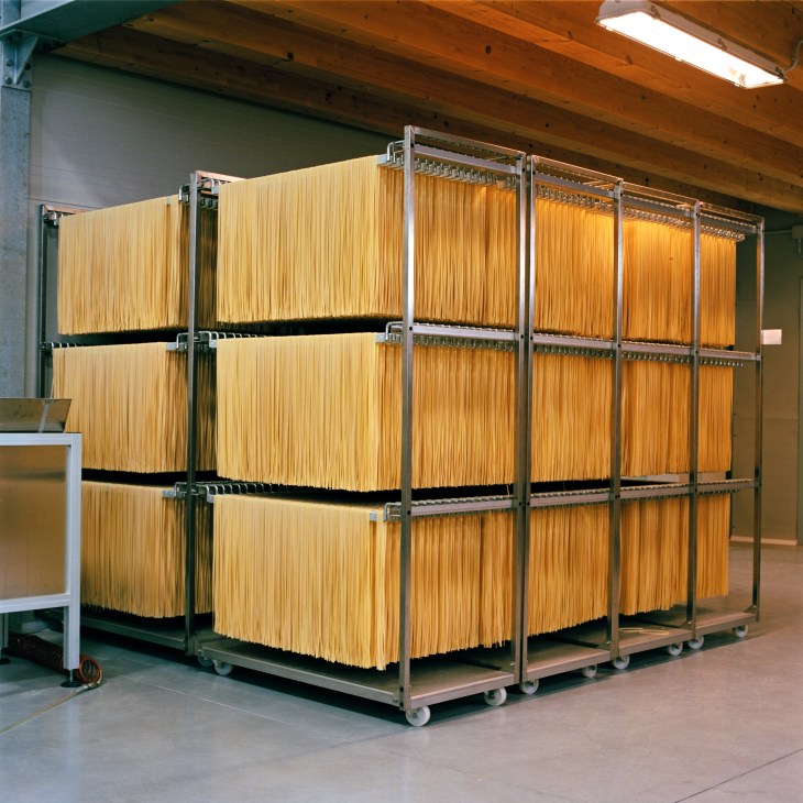 Industrial drying racks holding long strands of spaghetti in a factory with metal framework and wooden beams.