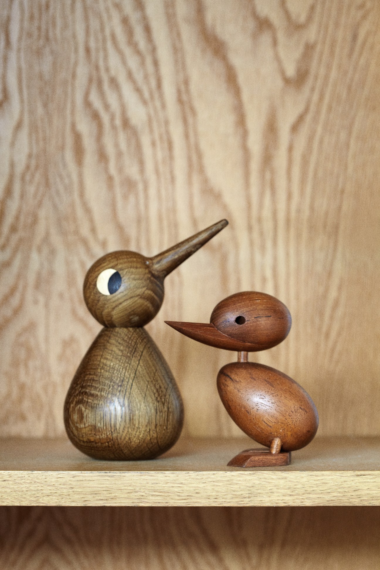 Two wooden bird sculptures on a shelf: a tall bird with a blue eye and a smaller duck-like figure.