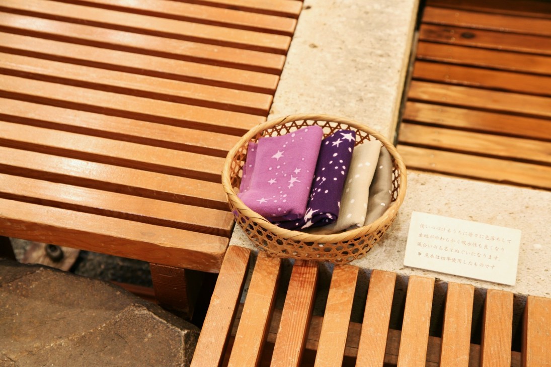 Hand-dyed tenugui cloths in purple, navy and cream folded in a woven basket on wooden steps.