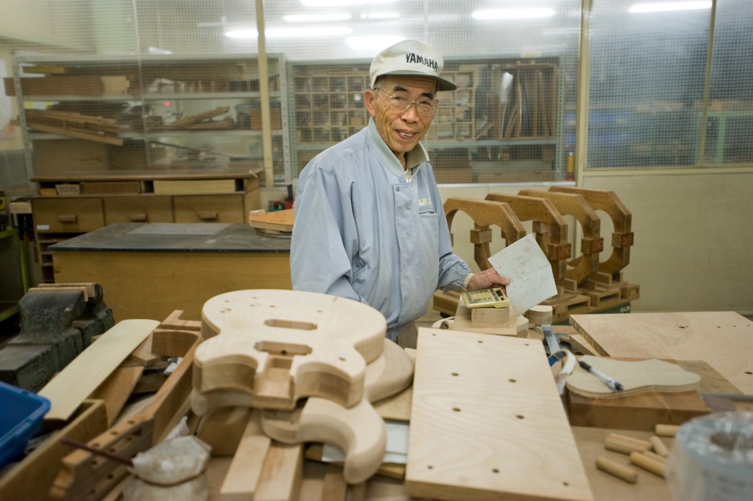 Tetsuya Samejima, Yamaha's experienced guitar maker, stands at a workbench with wooden guitar body parts and components.