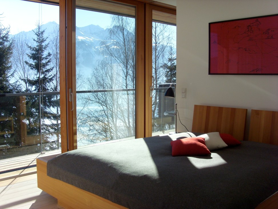 Minimalist bedroom with wooden bed frame, large windows overlooking snowy Alpine mountains and forest.