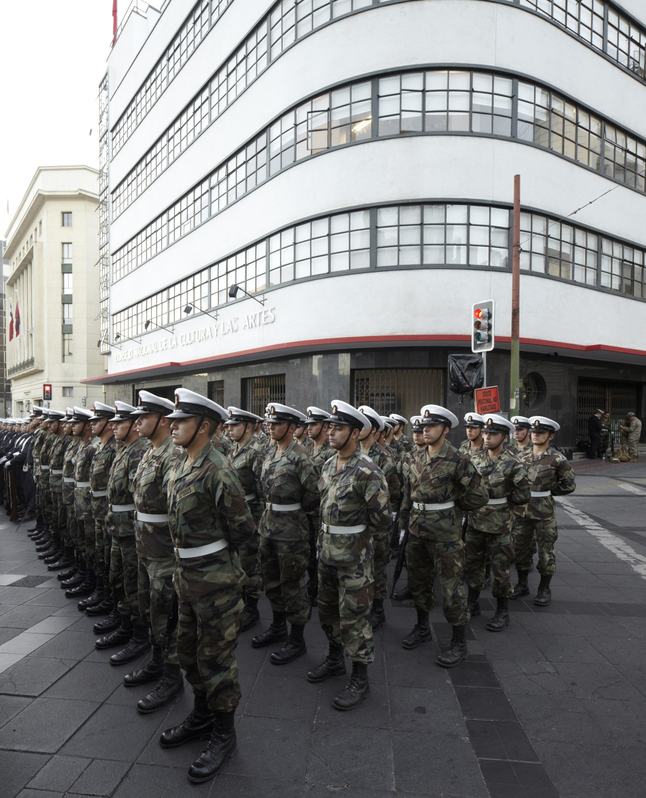Military personnel in camouflage uniforms and white naval caps stand in formation outside a modernist white building with curved windows.
