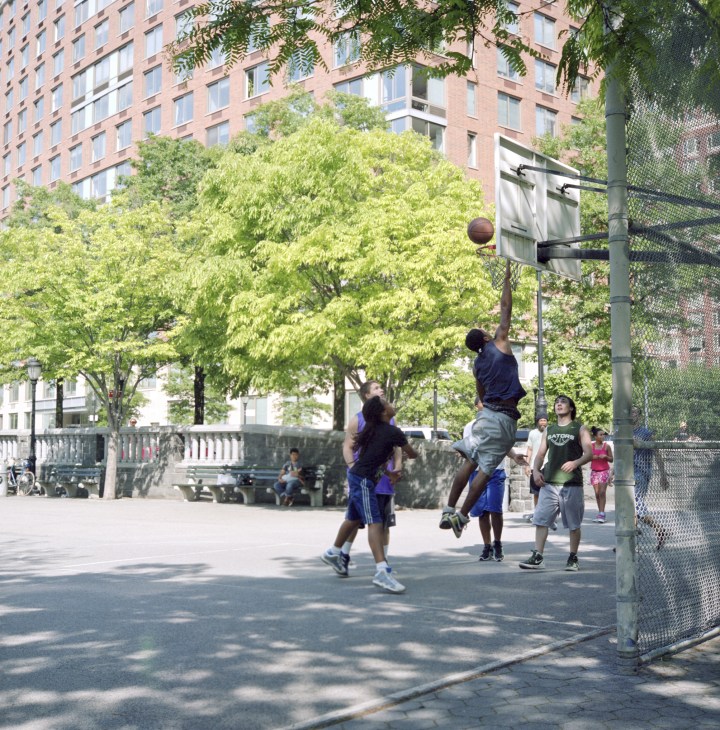 Children playing basketball on an outdoor court by the Hudson River with apartment buildings and green trees in the background.