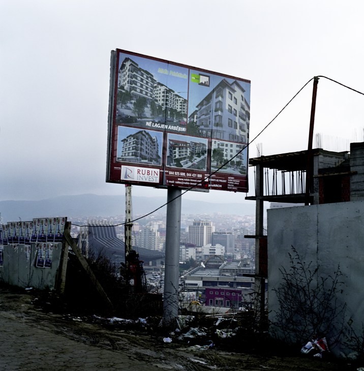 Real estate billboard advertising apartment buildings overlooking Pristina's cityscape with mountains in the background.