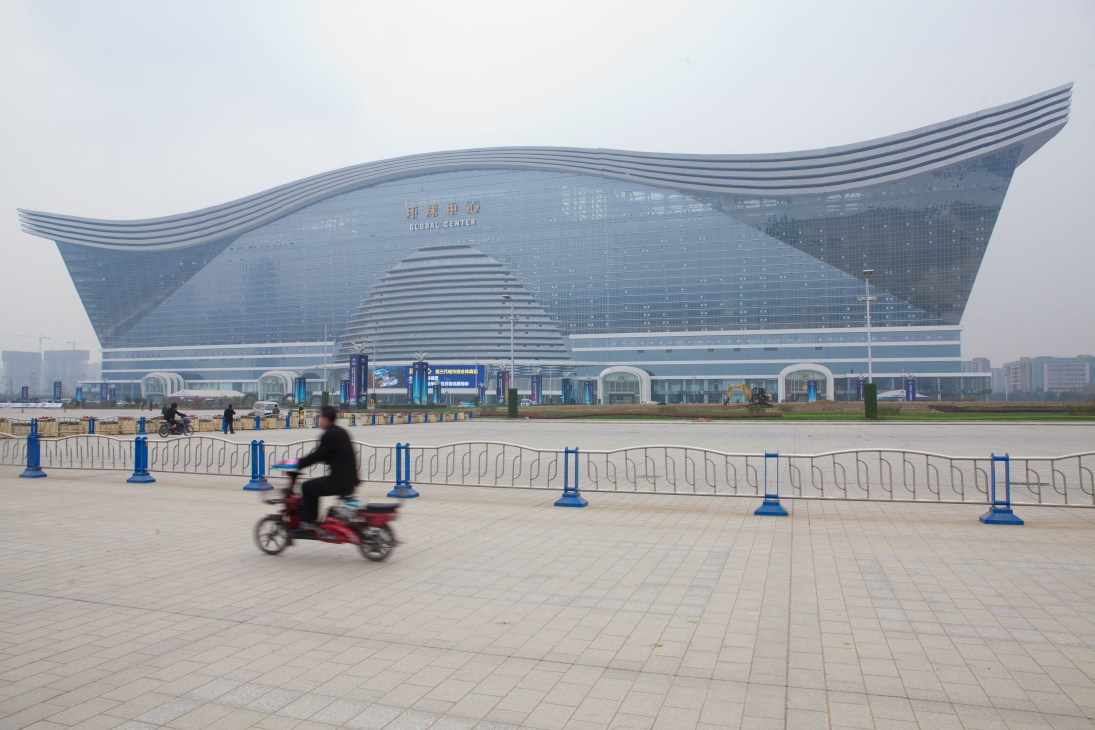 Large futuristic building with curved blue-tiled roof and person on red scooter in foreground.