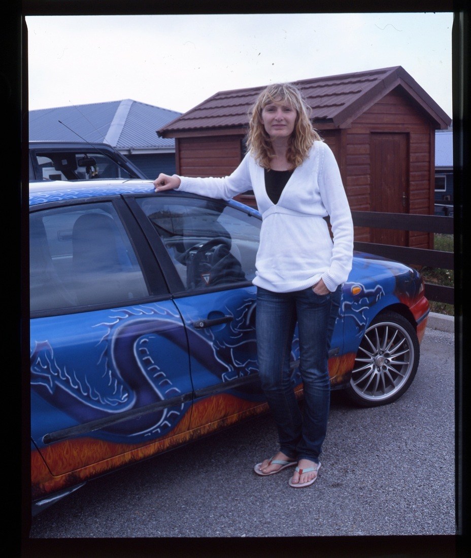A woman in a white cardigan stands beside a blue car with decorative paint in front of red garages.