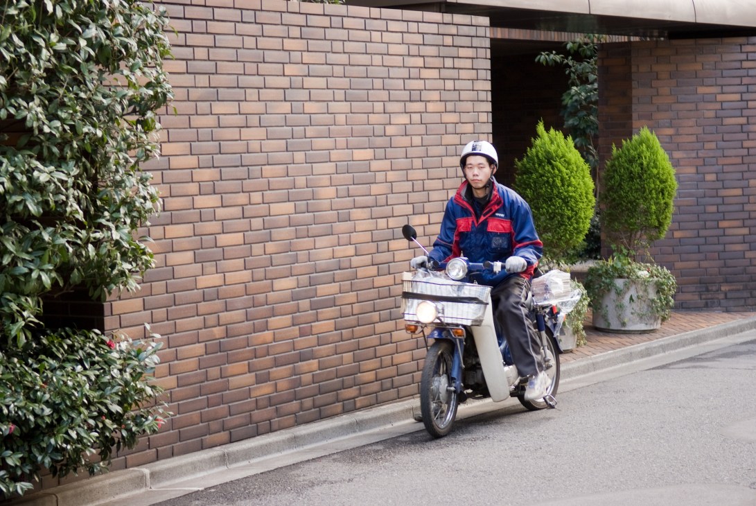 Newspaper delivery man riding a Honda Super Cub scooter past a brick building.