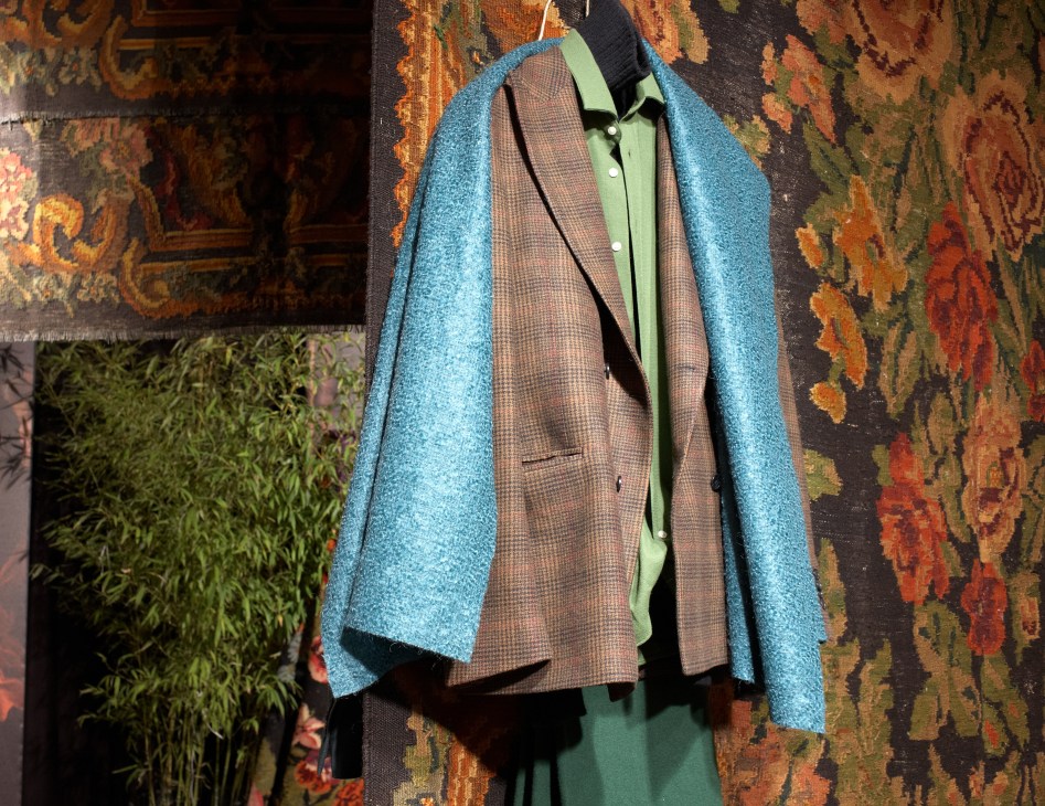 Colorful menswear display at Piombo featuring a turquoise jacket over a patterned blazer and green shirt against an ornate tapestry backdrop.