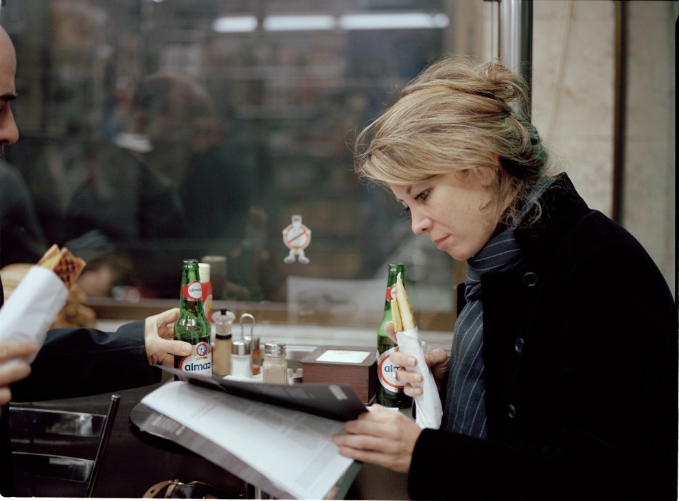 Woman with blonde hair in black pinstriped jacket holds Almaza beer bottle at table in restaurant