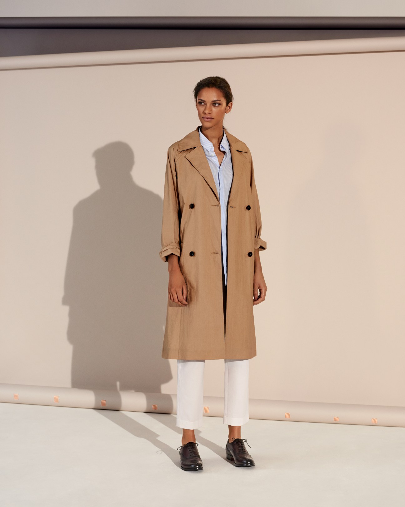Model wearing a beige double-breasted coat over a light blue shirt with white trousers and black shoes.