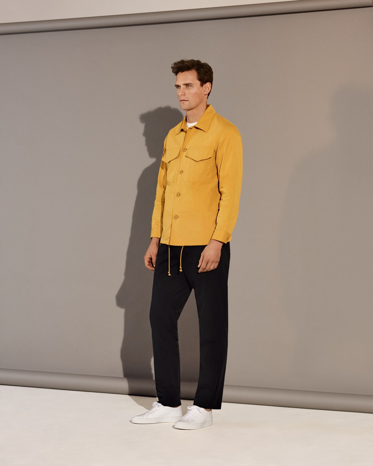Man wearing yellow jacket by Tone, black trousers by Acne Studios, and white trainers by Common Projects.