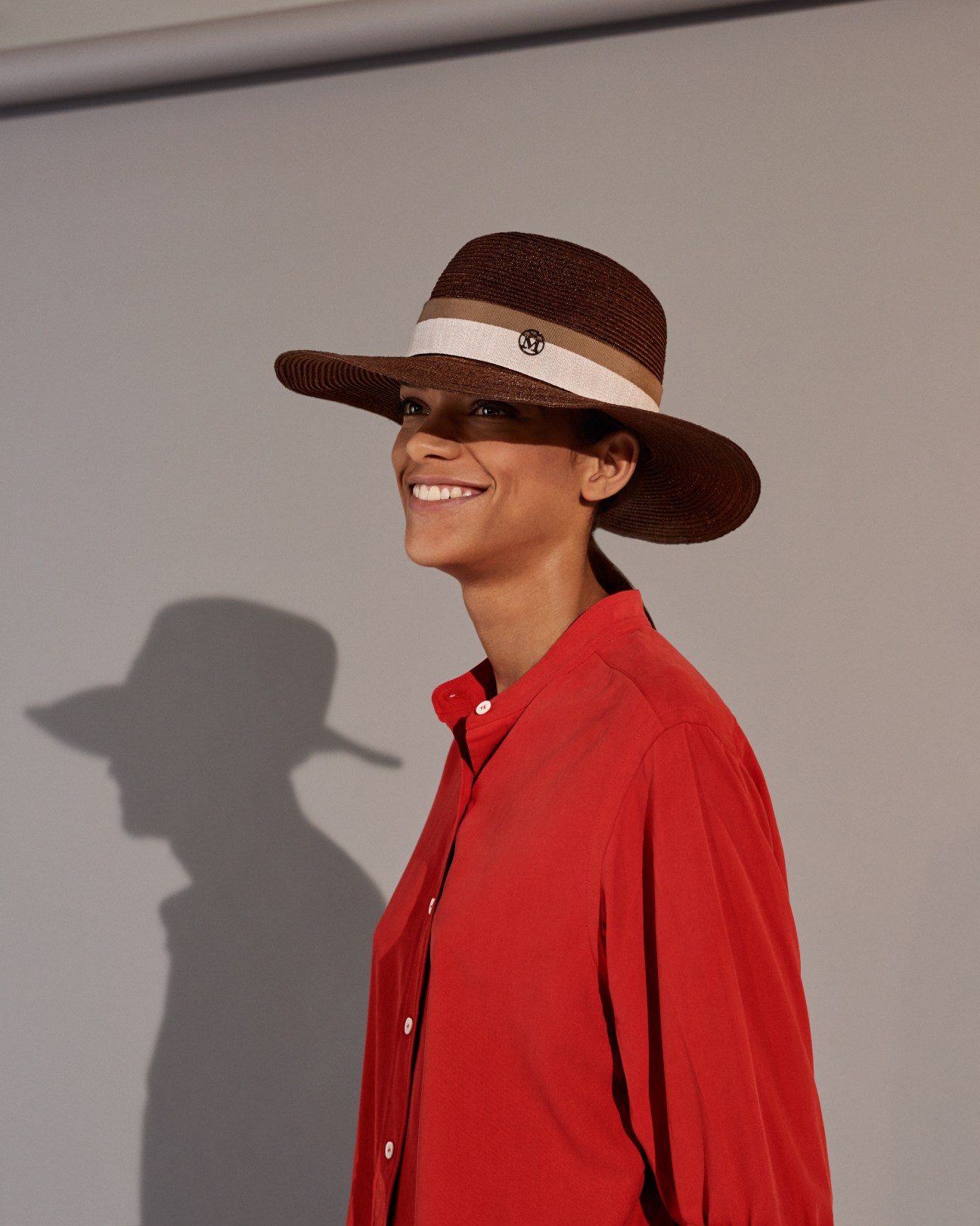 Woman wearing a red Holland & Holland shirt and brown Maison Michel wide-brimmed hat with cream band.