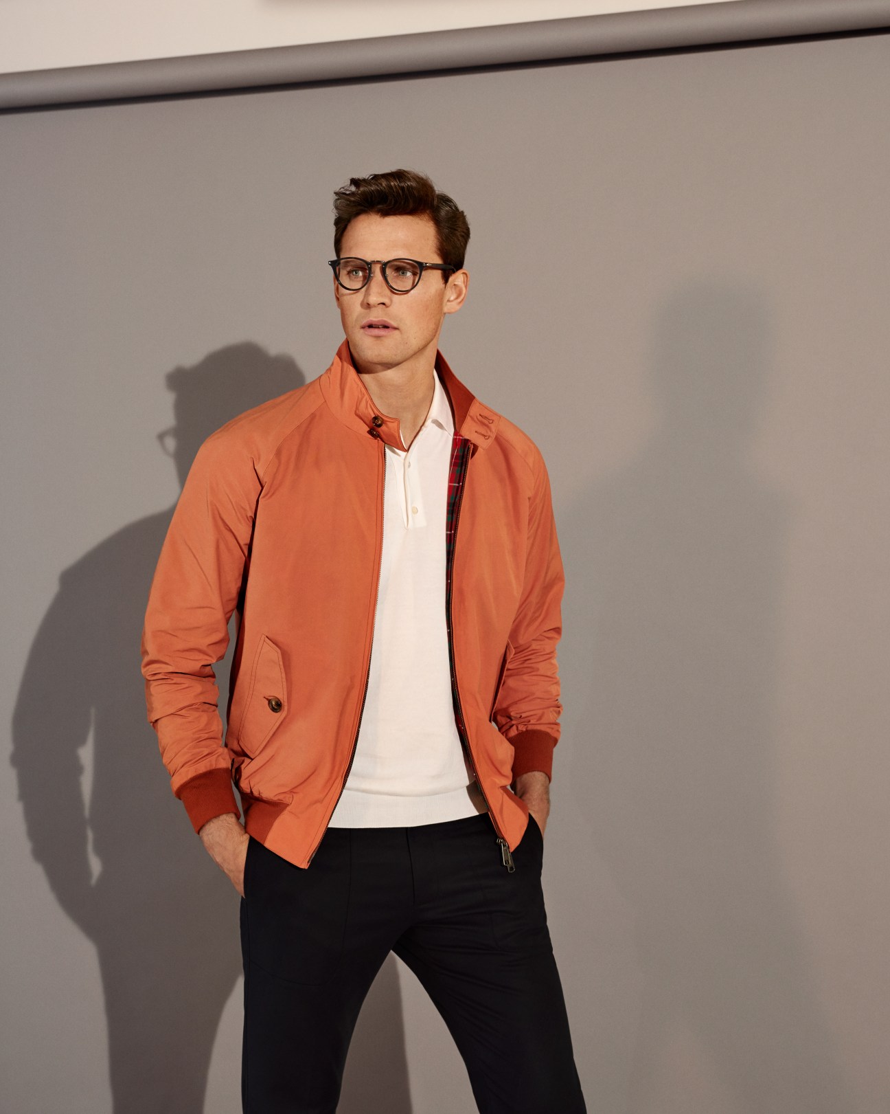 Man wearing orange Baracuta blouson, white polo shirt, black trousers and Persol glasses against grey background.