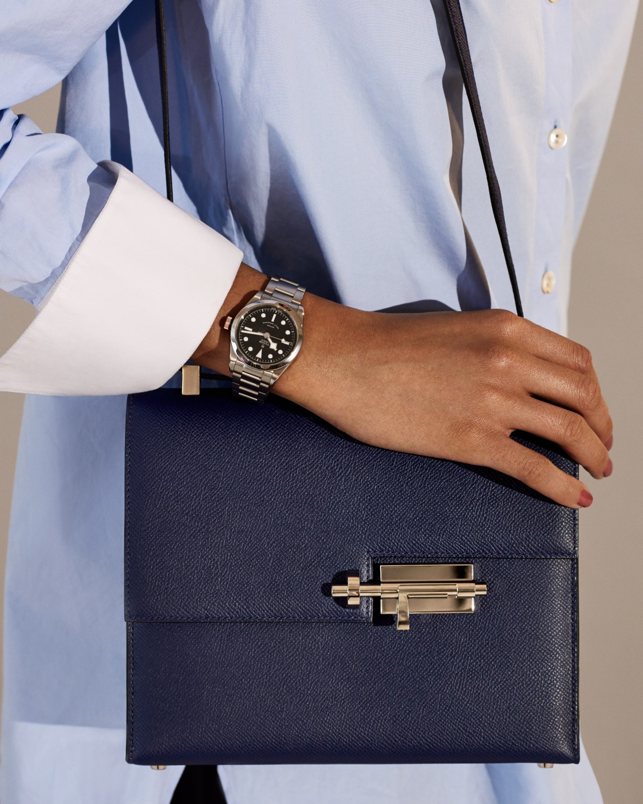 Person wearing light blue shirt holding navy Hermès bag with Tudor watch on wrist.