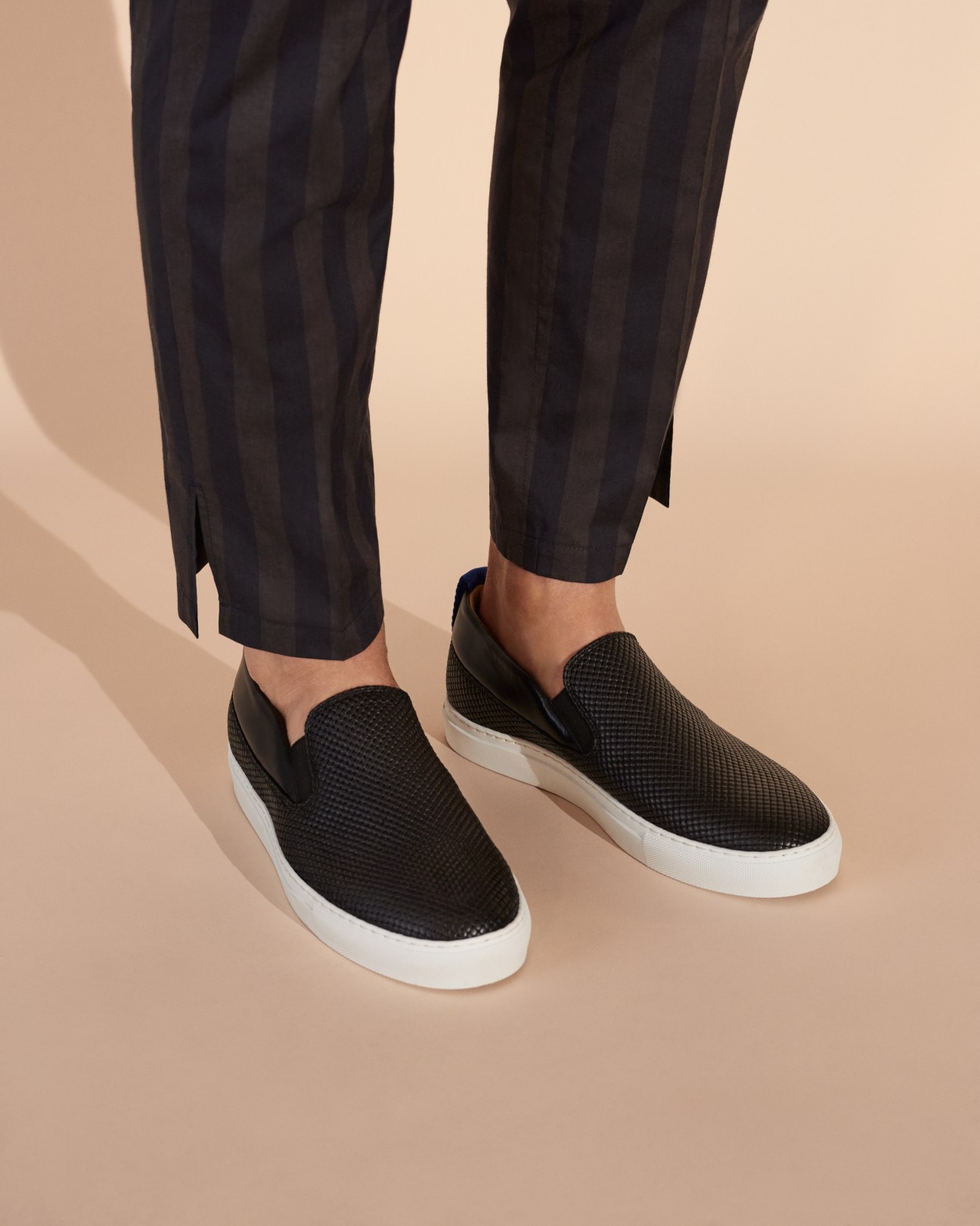 Black slip-on sneakers with white sole worn with dark striped trousers on beige background.