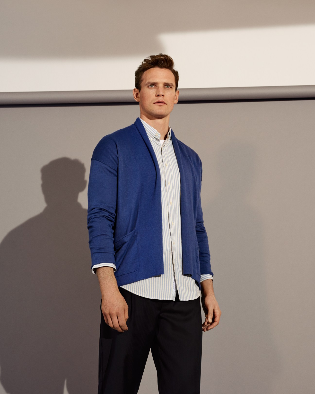 Man wearing a blue cardigan by Blue Work, white striped shirt, and black trousers against a minimalist background.
