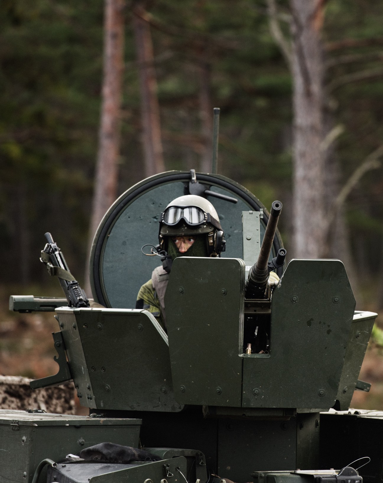 Swedish soldier in protective gear operating a mounted machine gun from a fortified military position in a forest.