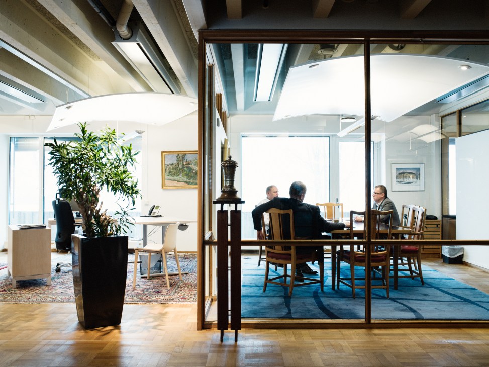 Three businessmen in a glass-walled meeting room at Scania headquarters with modern furnishings and potted plants visible.