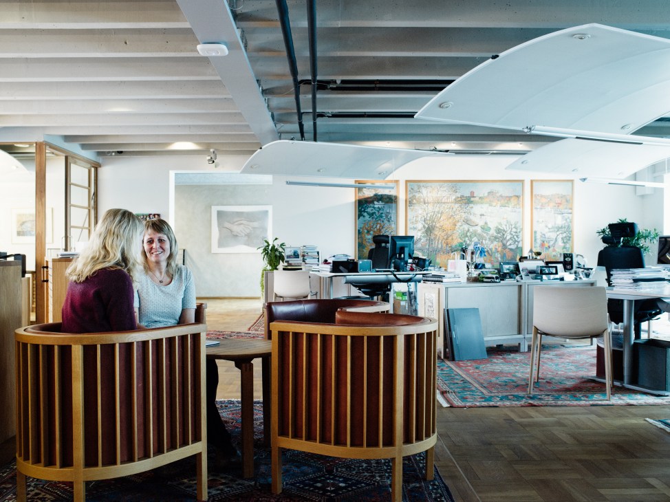 Two women seated at round wooden mid-century desks in a bright office with Persian rugs and artwork on walls.