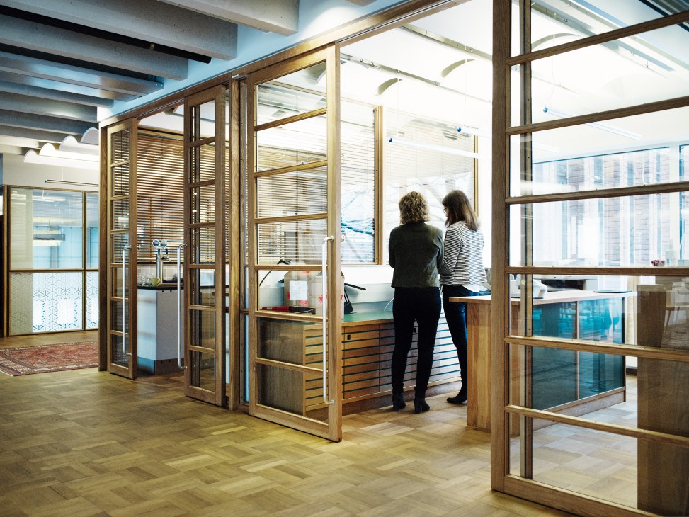 Two women stand inside a glass-walled office space with wooden frames and horizontal louvers at Scania headquarters.