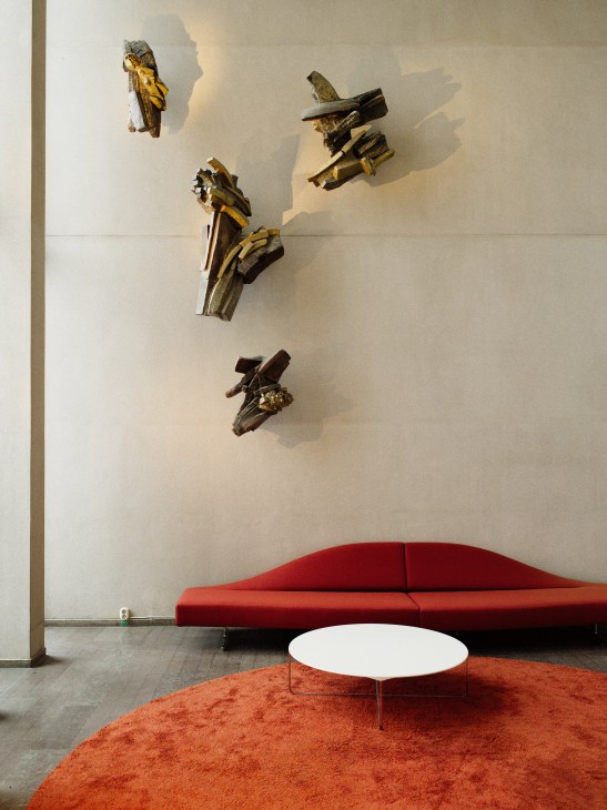 Modern lobby with red curved sofa, white round table on orange rug, and abstract wooden sculptures on white wall.
