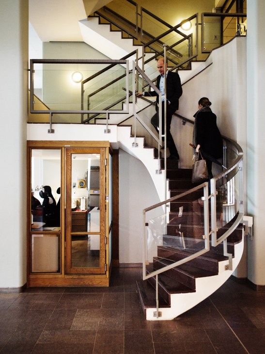 A modern spiral staircase with black metal railings and wooden handrail inside Scania's office building, with employees walking up the stairs.