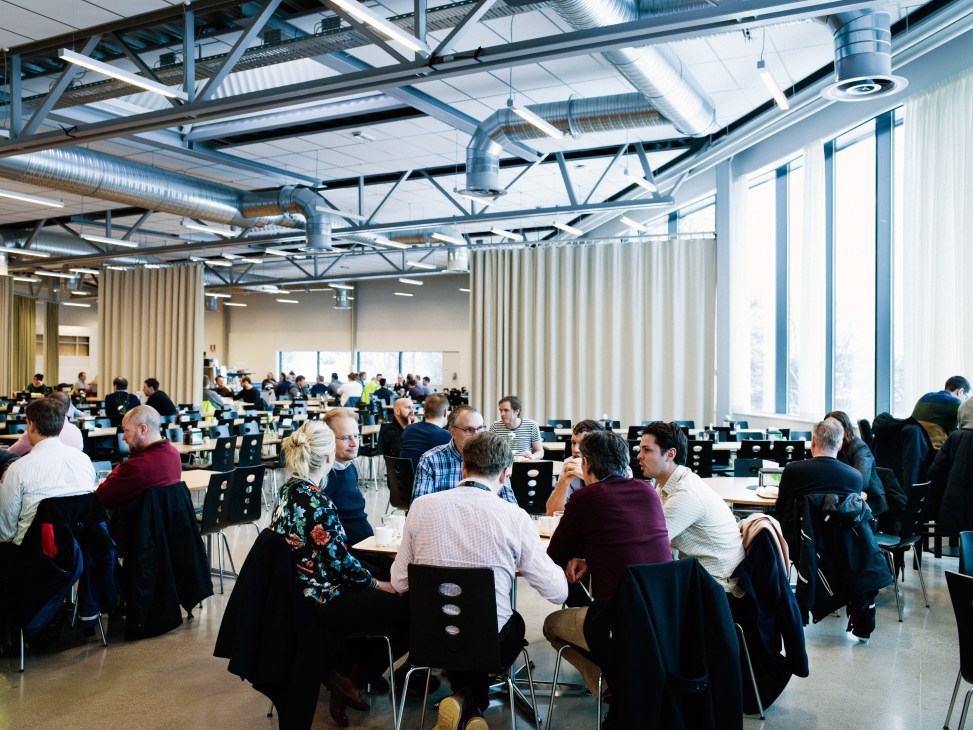A large industrial canteen with steel beams and white curtains where Scania R&D employees dine at tables.