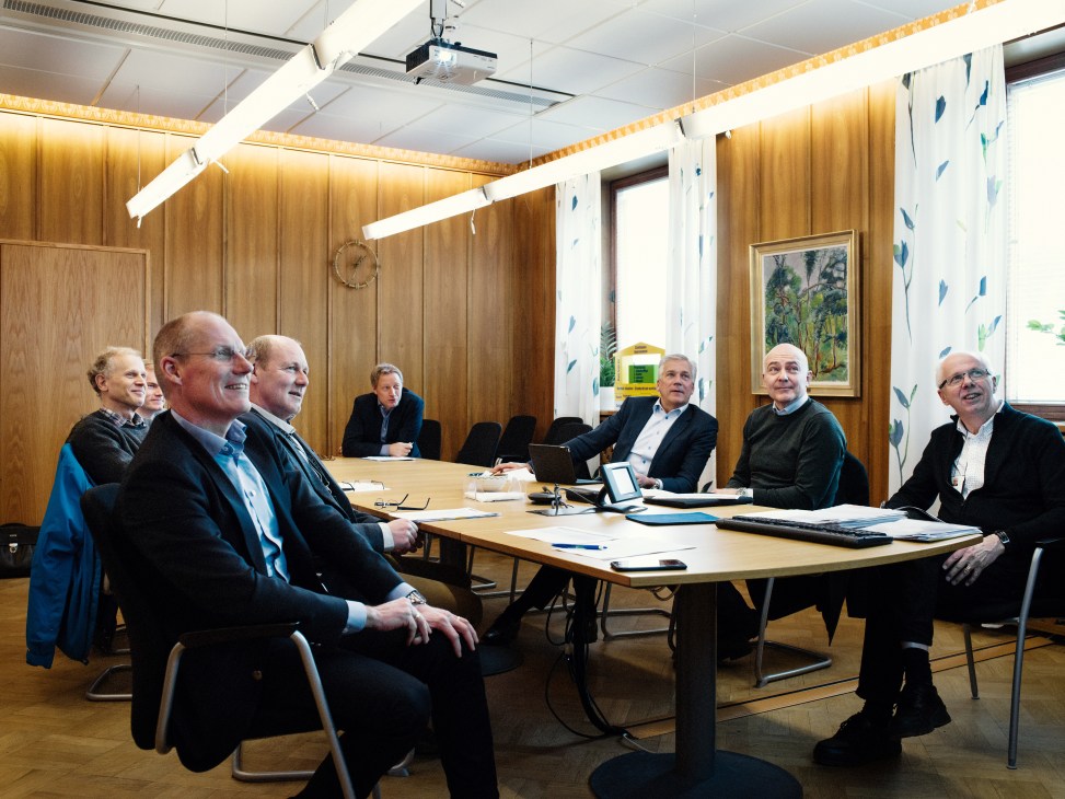 Scania employees seated at a wooden table in a wood-paneled meeting room with blue patterned curtains.