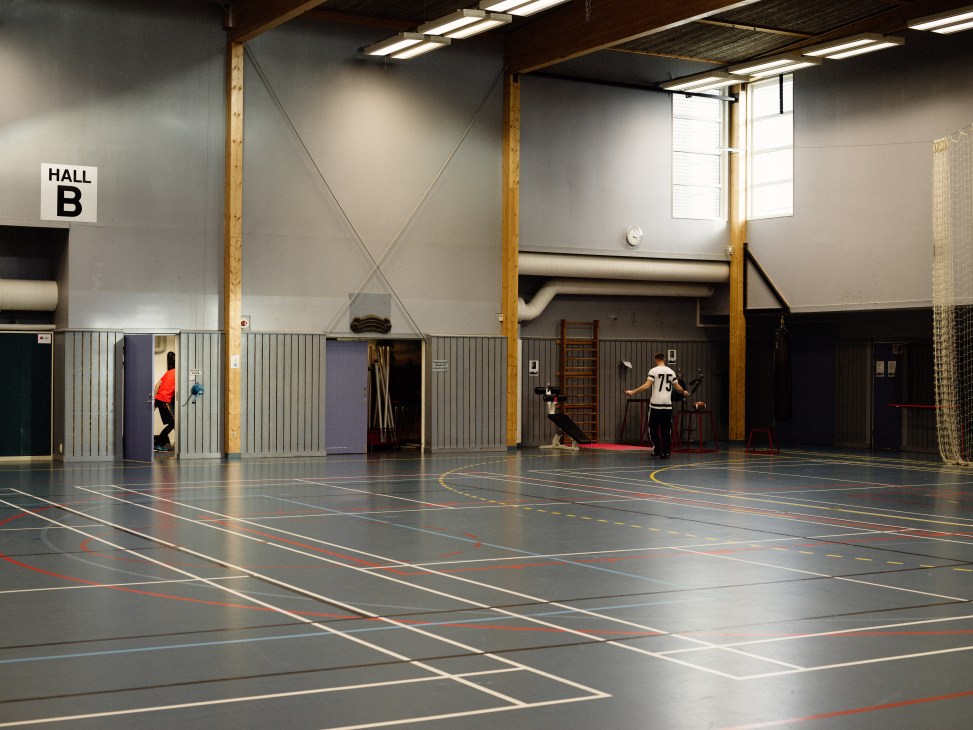 Two people exercising in a large modern sports hall with wooden support columns and marked court lines.