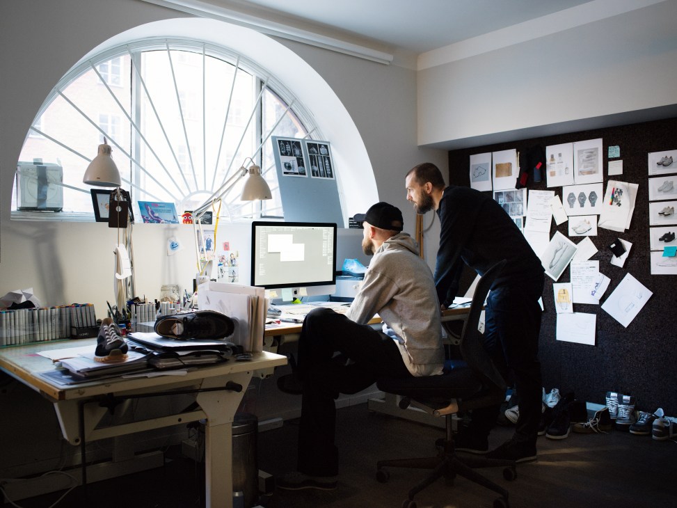 Two men work in a bright studio with a large round window, surrounded by design sketches and shoe samples on desk and walls.