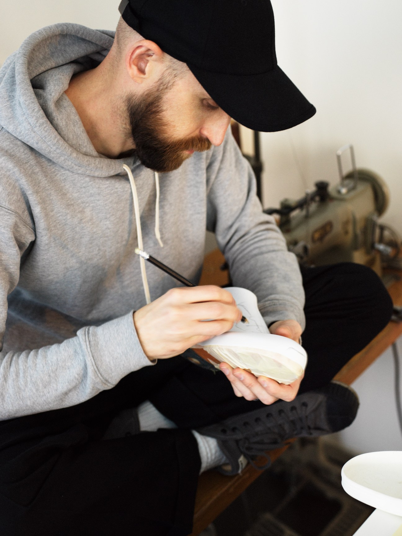 Man in gray hoodie and black cap examining a white shoe mould in his hands.