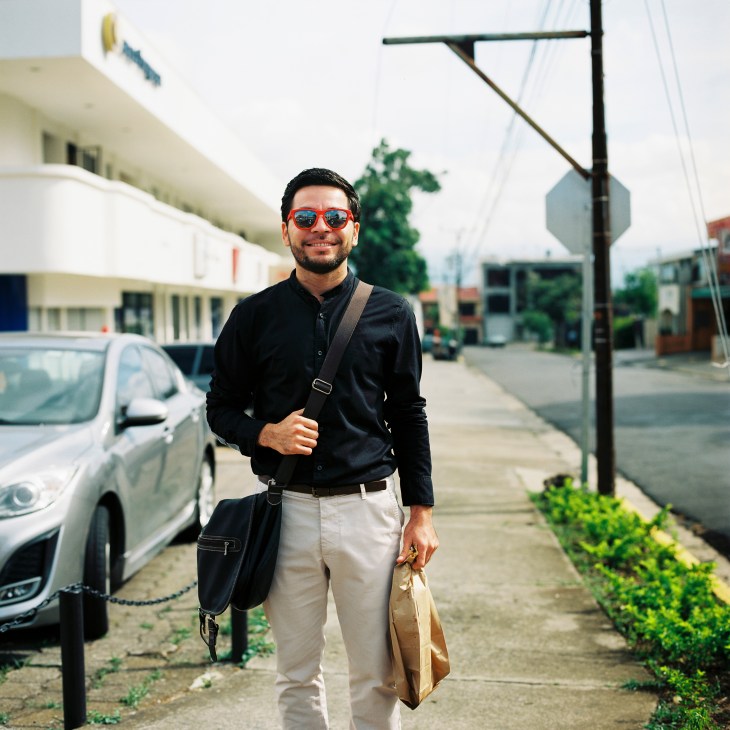 Man in black shirt and sunglasses walks down a San José street carrying a tan bag.