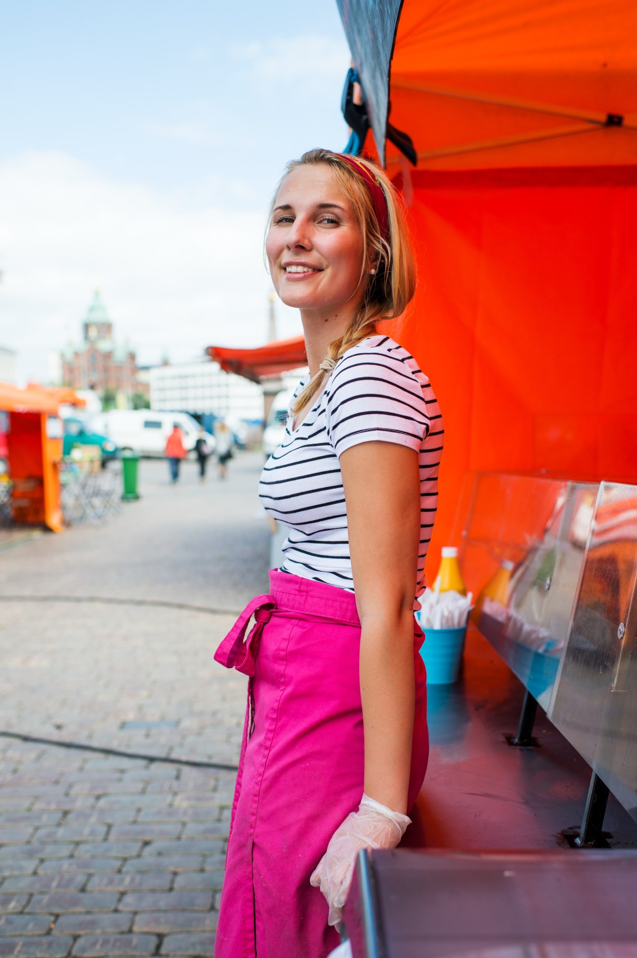 Woman in striped shirt and pink pants standing by red food truck in Helsinki with cathedral in background.