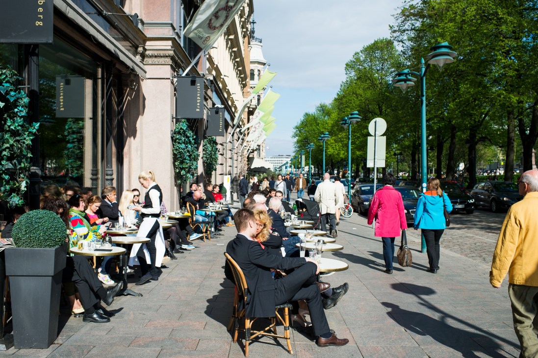 Outdoor street dining scene in Helsinki with people seated at tables along a tree-lined pedestrian avenue.