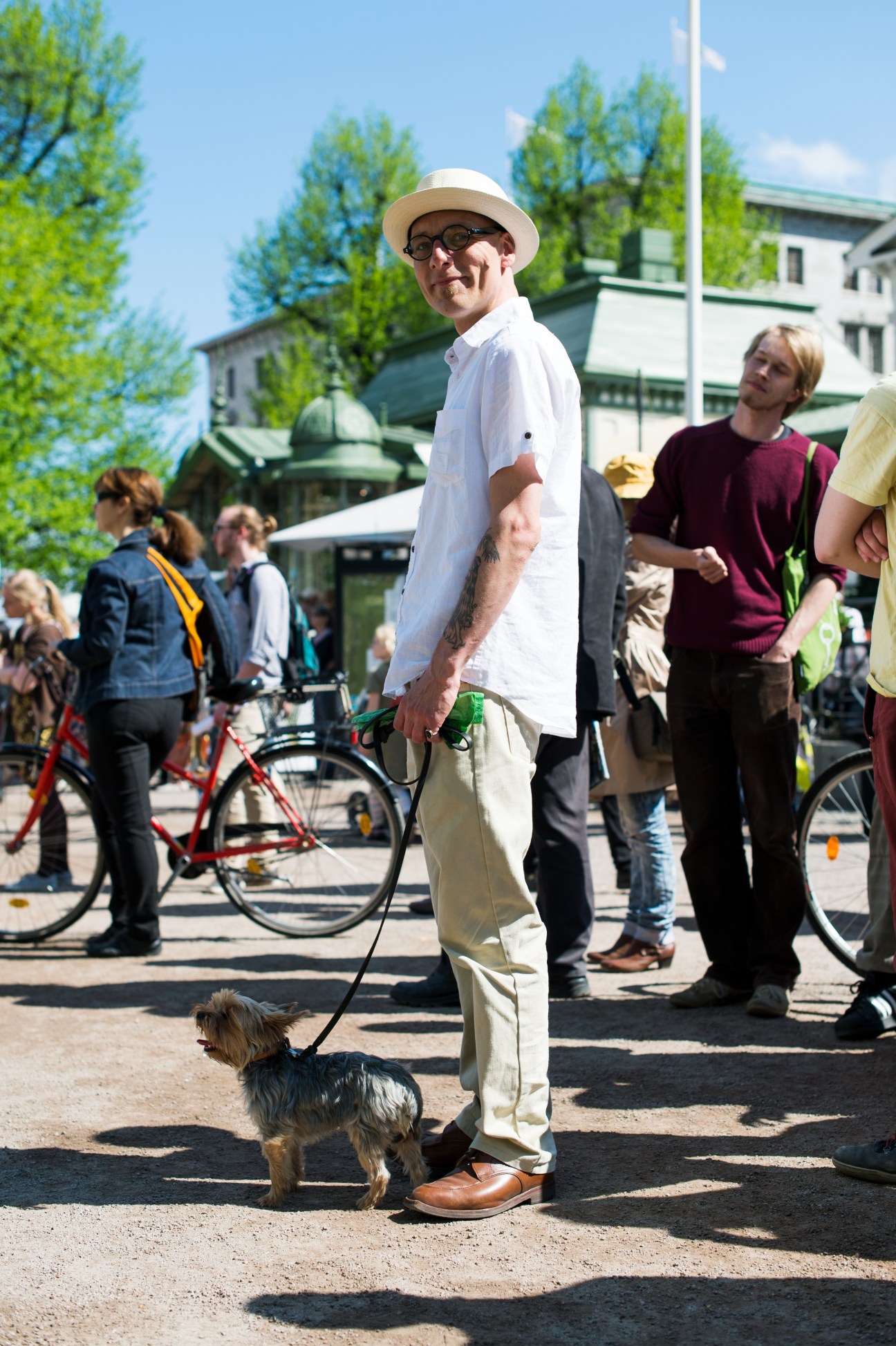 Man in white shirt and Panama hat walks small dog through Helsinki street with cyclists and pedestrians.