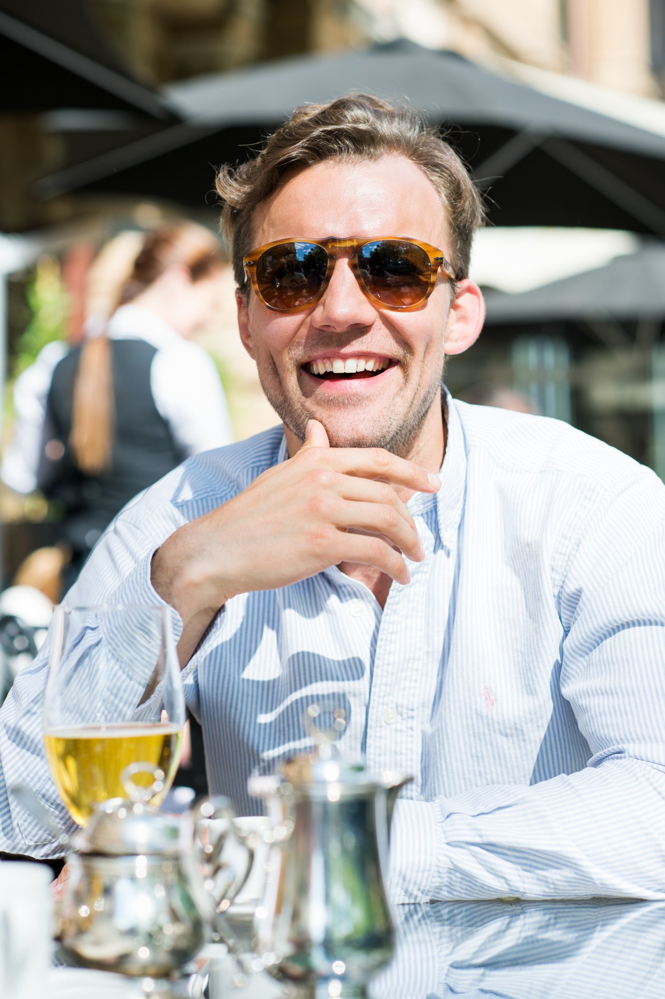 Man wearing sunglasses and blue shirt smiling at outdoor café table with beer and water glasses.