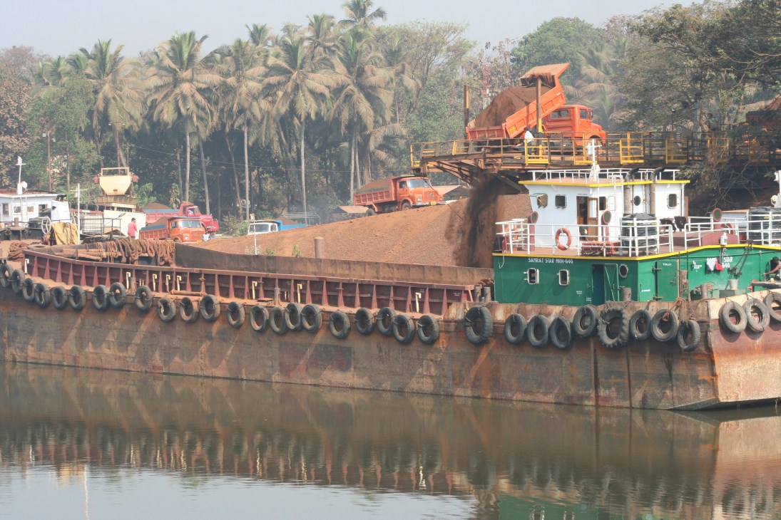 Iron ore being loaded onto a cargo ship in Goa, India with mining trucks and palm trees visible onshore.
