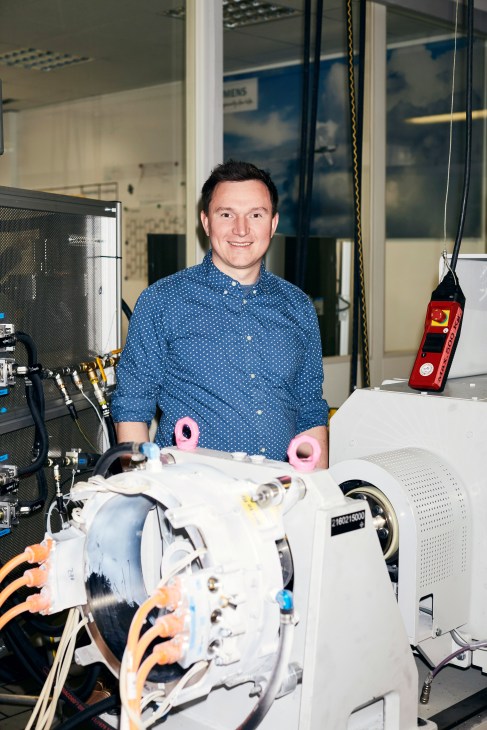 Man in blue polka dot shirt smiling beside compact electric motor with wires and sensors in laboratory setting.