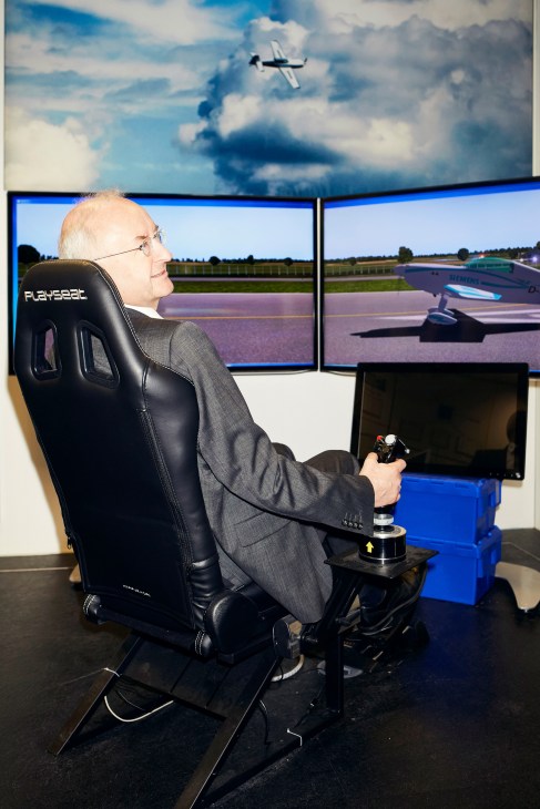 Frank Anton, head of Siemens eAircraft, sits in a racing simulator chair holding a joystick while viewing screens displaying a virtual aircraft and runway.