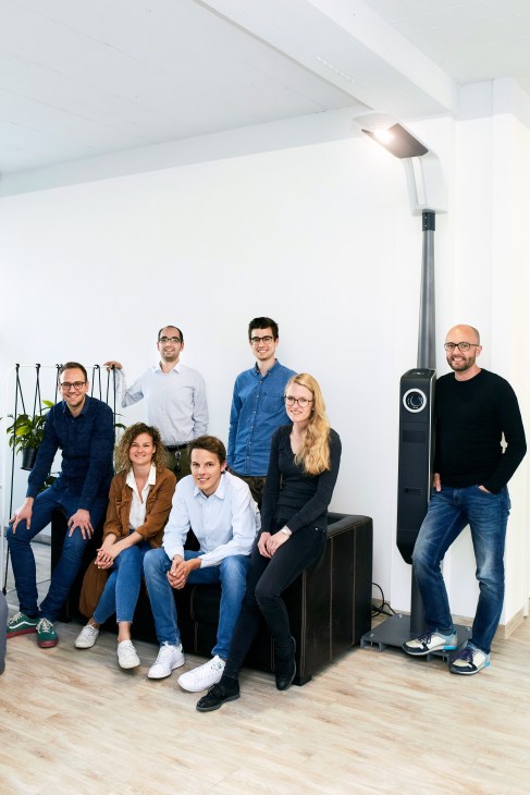 Eluminocity team of eight people posing in a bright white office space with plants and modern lighting.