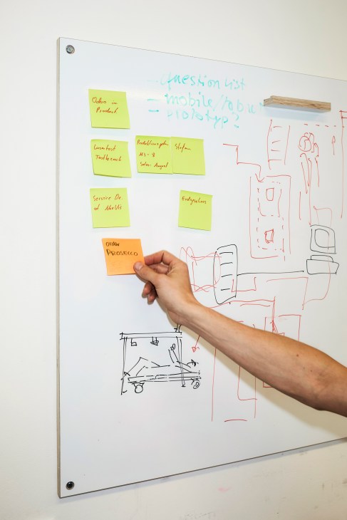 A person's hand places an orange sticky note on a whiteboard covered with yellow notes, sketches and handwritten text.