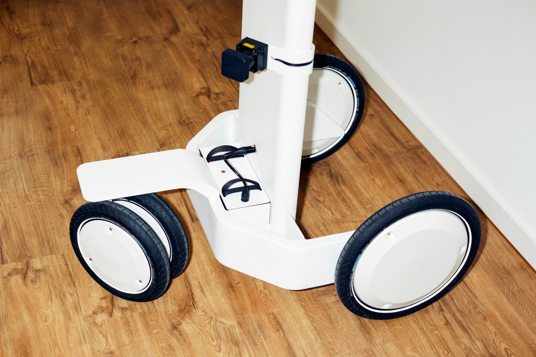 Close-up of a white wheeled device with black tires and sensor box, showing battery and wheel components on wooden floor.