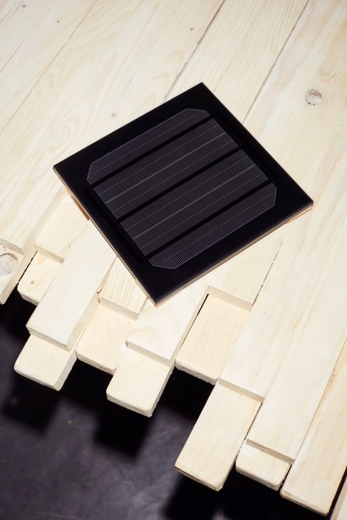 Solar panel with fine line pattern mounted on wooden blocks on light wood surface.