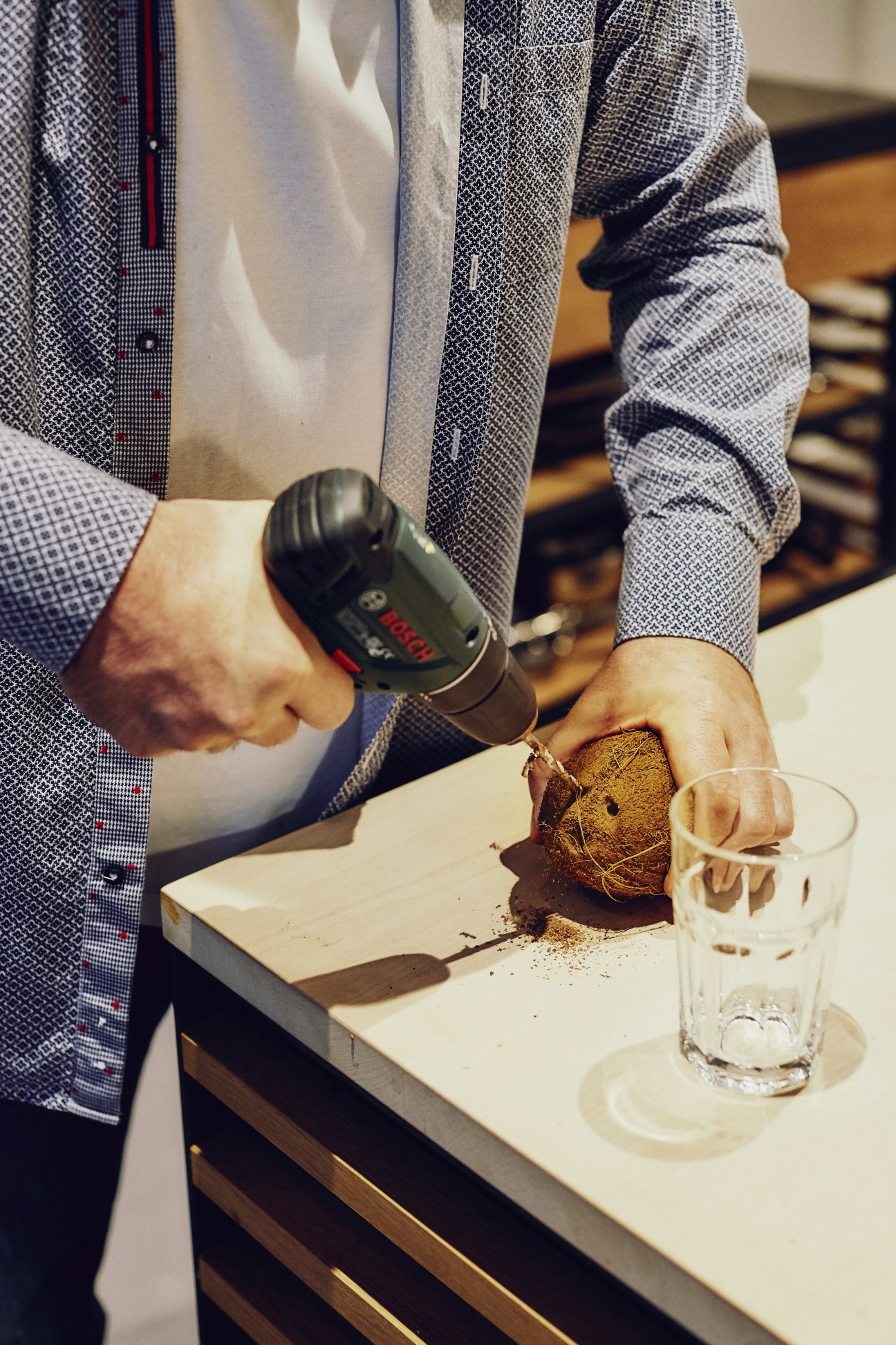 Person using a Bosch drill to extract coconut water from a halved coconut on a white surface.